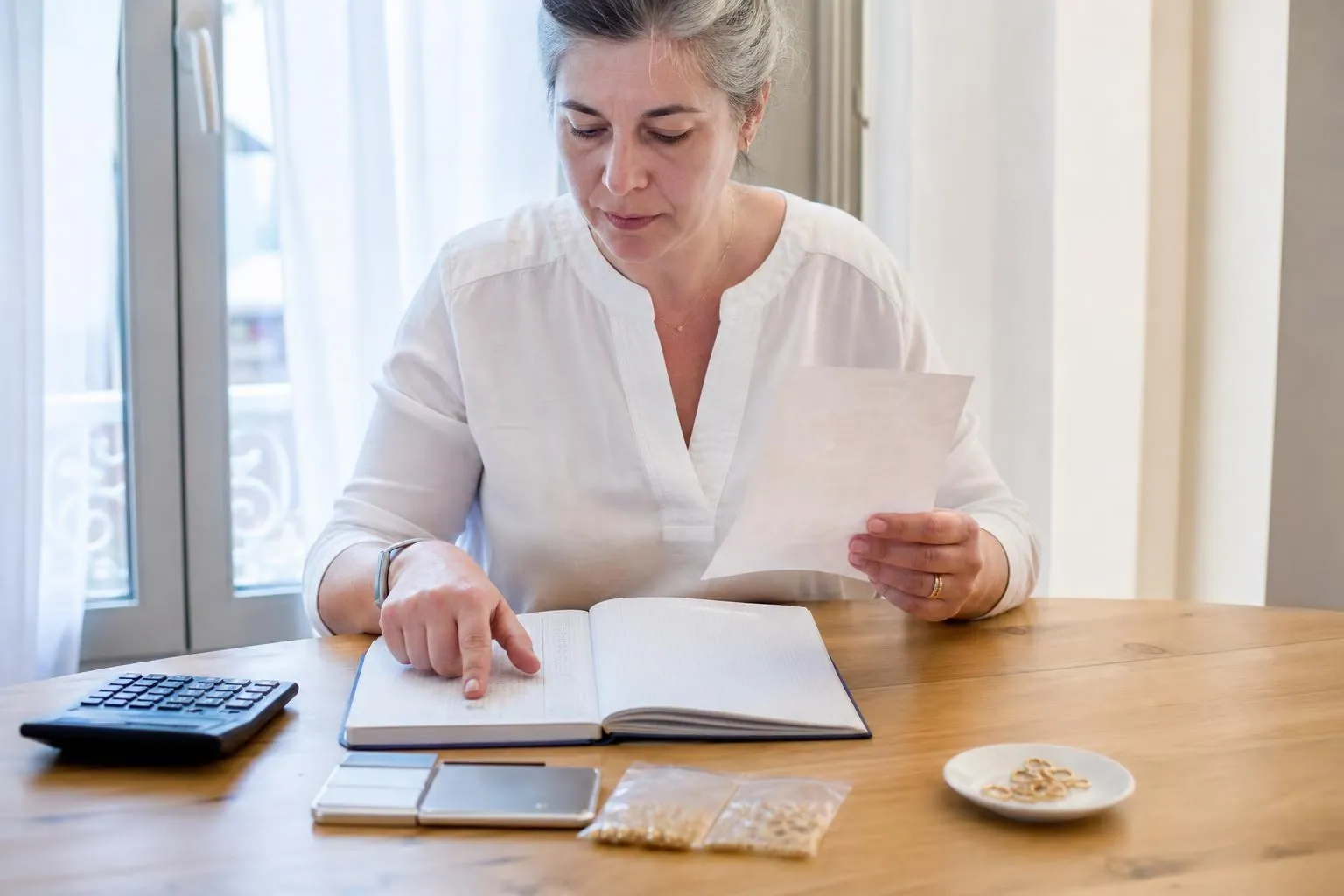 Créatrice comparant des factures d’apprêts et un cahier de comptes sur une table claire, avec une calculatrice et une balance de précision