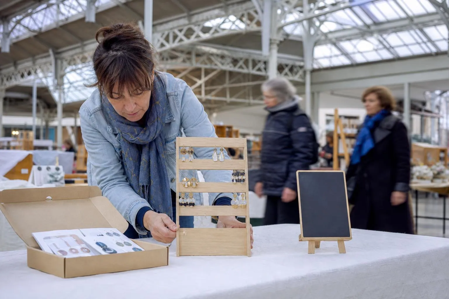Créatrice disposant ses présentoirs de boucles et une pochette de fiches sur un stand de marché de créateurs couvert